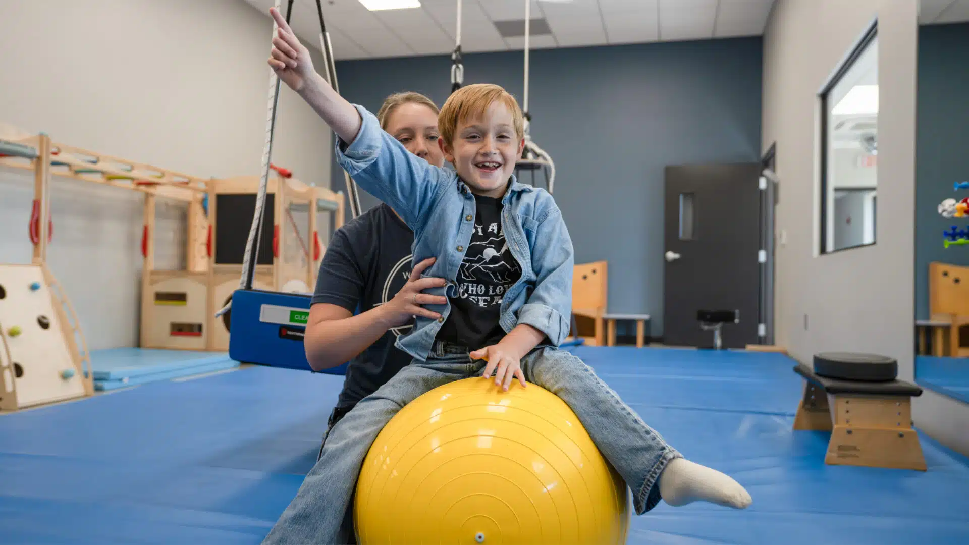A little boy sitting on an exercise ball with assistance from a physical therapist.